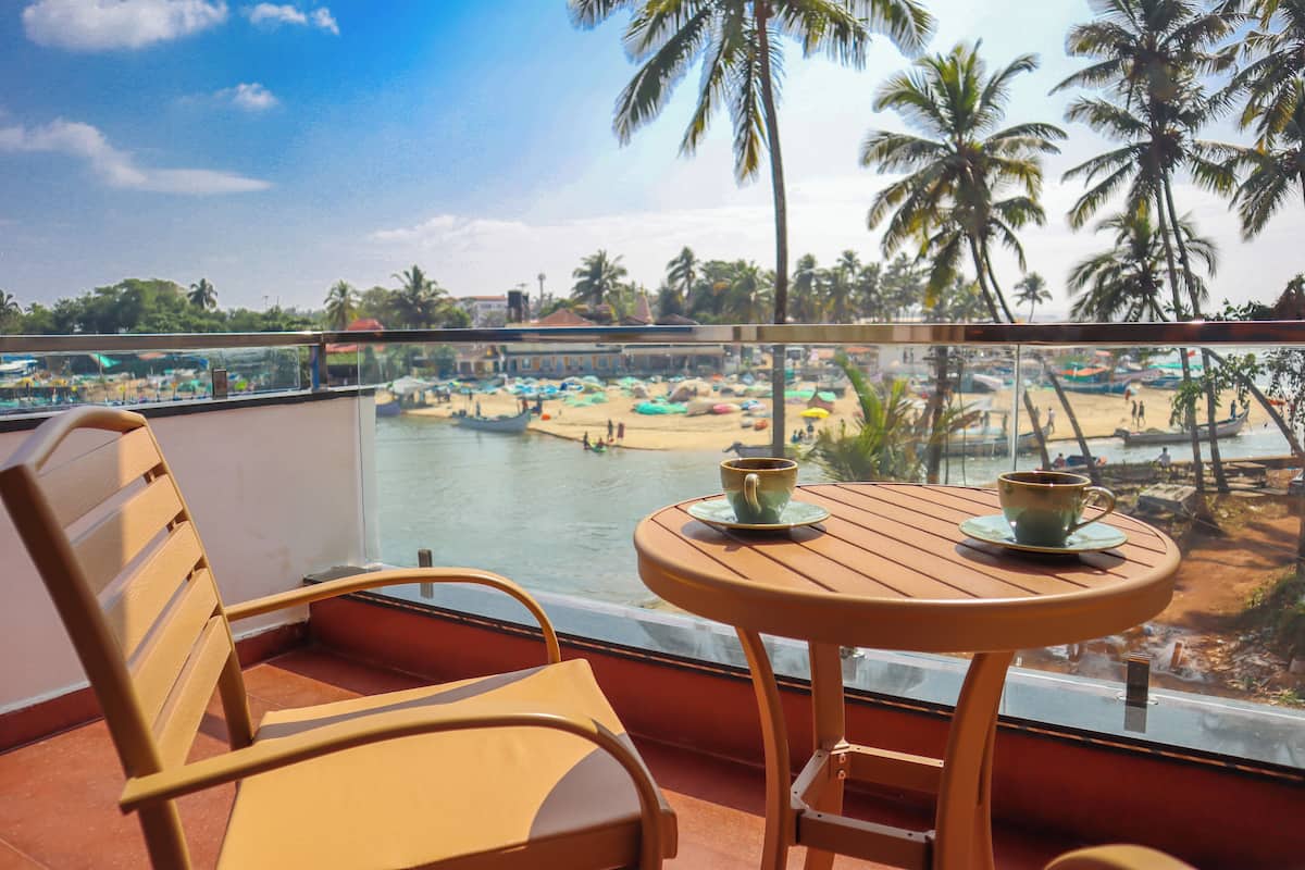 Un balcón con una silla y mesa de madera, con vista a una playa con barcos y palmeras.