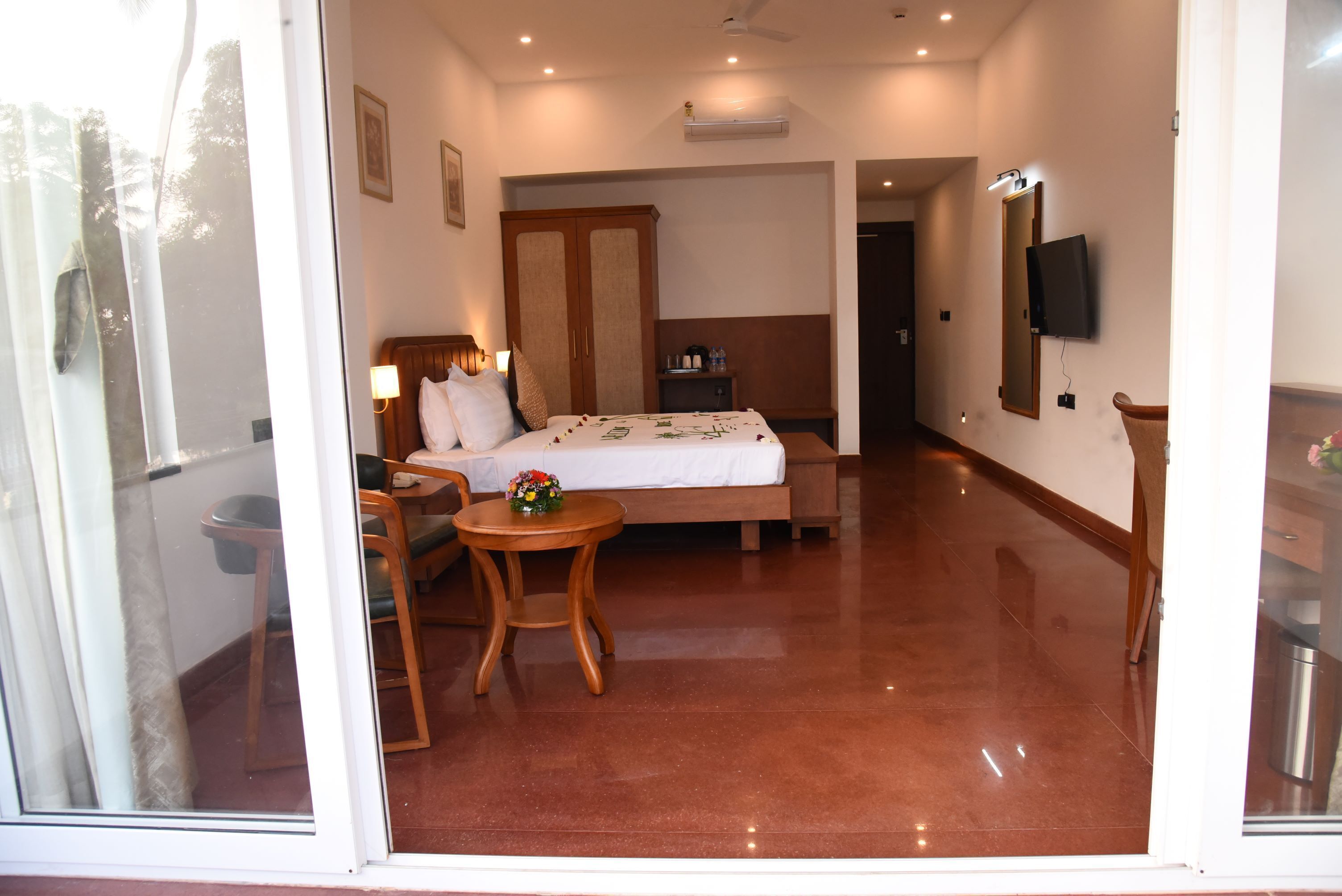 A hotel room with a bed, a desk, a chair, a small table with a flower, and a TV mounted on the wall.