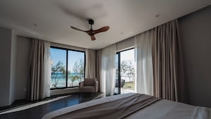 A modern hotel room with a large bed, a ceiling fan, and a view of the sea through large windows.