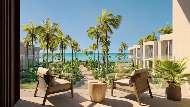 A balcony with two wicker chairs and a round table, overlooking a beach with palm trees and a clear blue sky.