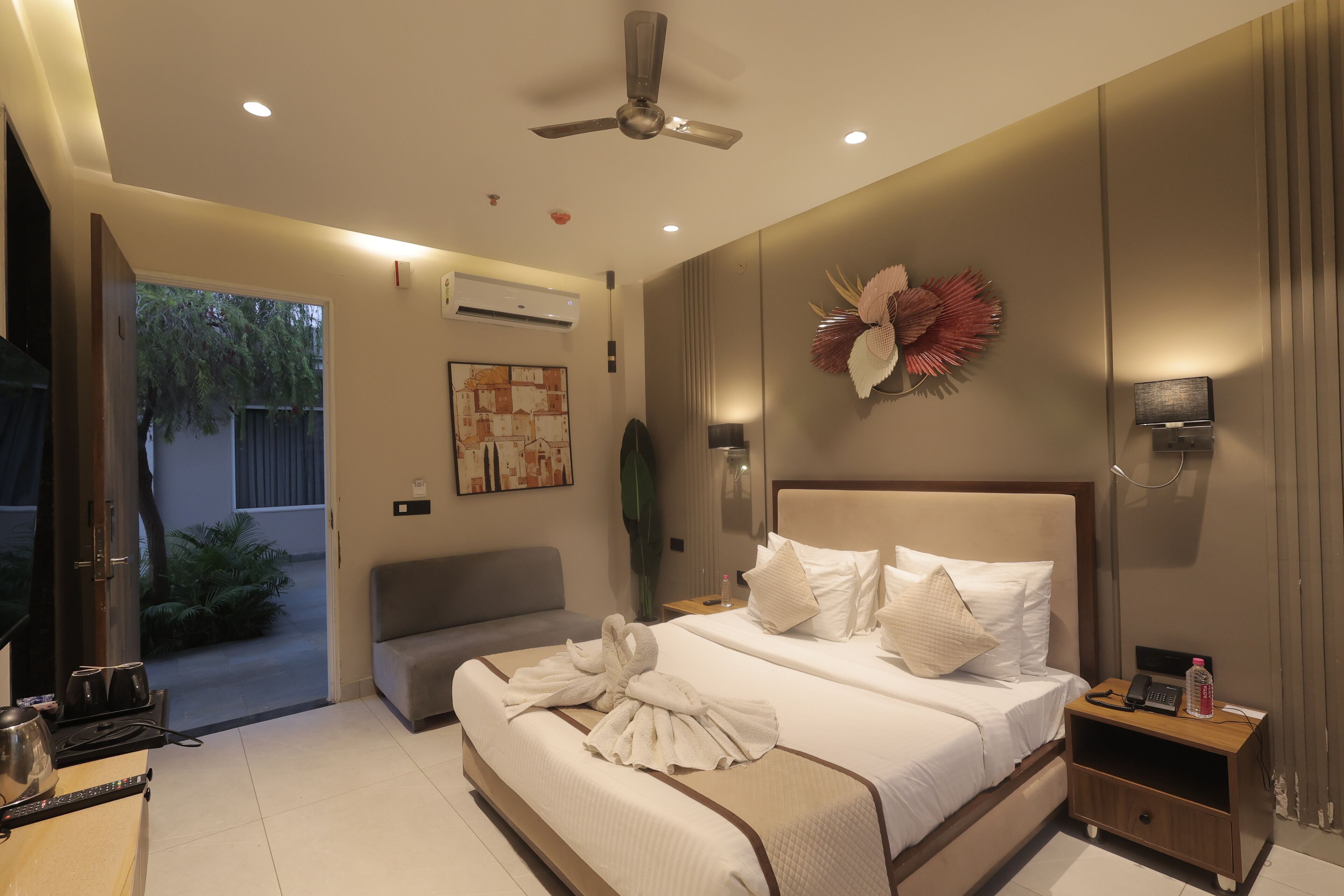 A modern hotel room with a large bed, a bedside table with a phone and a bottle, a ceiling fan, and a view of the outside.