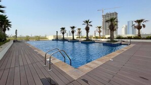 A rooftop swimming pool with palm trees and a view of a cityscape with high-rise buildings. - Bespoke Residences - Tower 108 JVC (Dubai)