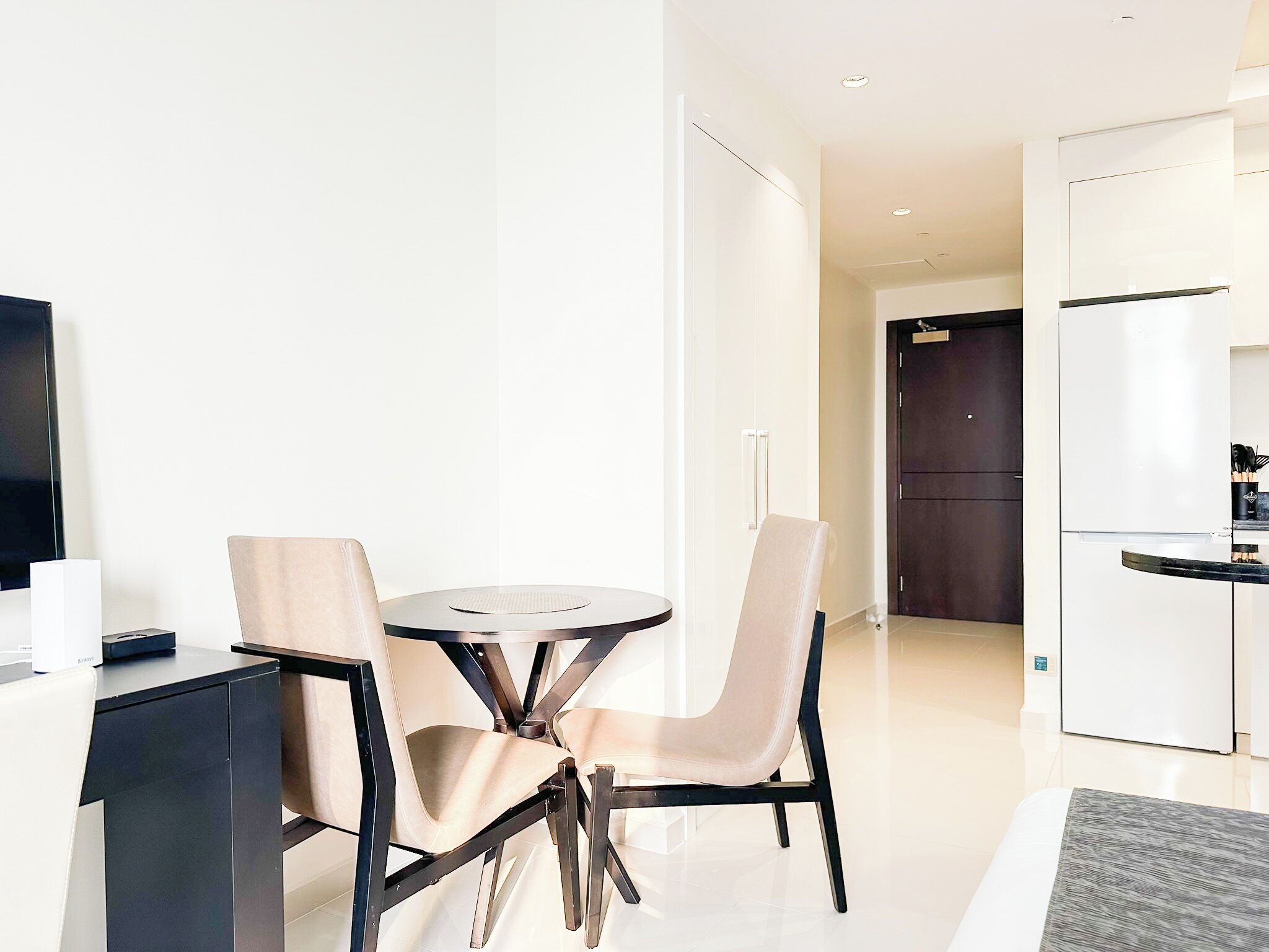 A modern hotel room with a dining table and chairs, a flat-screen TV, a refrigerator, and a dark wooden door.