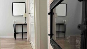 Un couloir doté d’un plancher en bois, d’un miroir encadré de noir accroché au mur et d’une petite table sur laquelle se trouve une lampe.