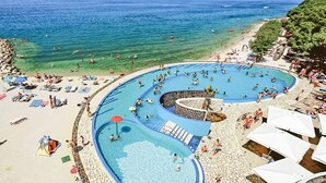 Une grande piscine extérieure divisée en plusieurs zones, entourée d’une plage de sable et offrant une vue dégagée sur la mer.