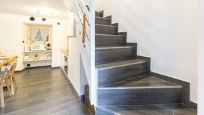 Un intérieur moderne avec un escalier menant à un deuxième étage, un coin repas avec des chaises en bois et une cuisine dotée de meubles blancs et d’un four à micro-ondes.