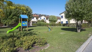 Un terrain de jeux avec un toboggan vert et une structure d’escalade bleue, entouré d’une pelouse et d’arbres bien entretenus, avec des immeubles d’habitation en arrière-plan.