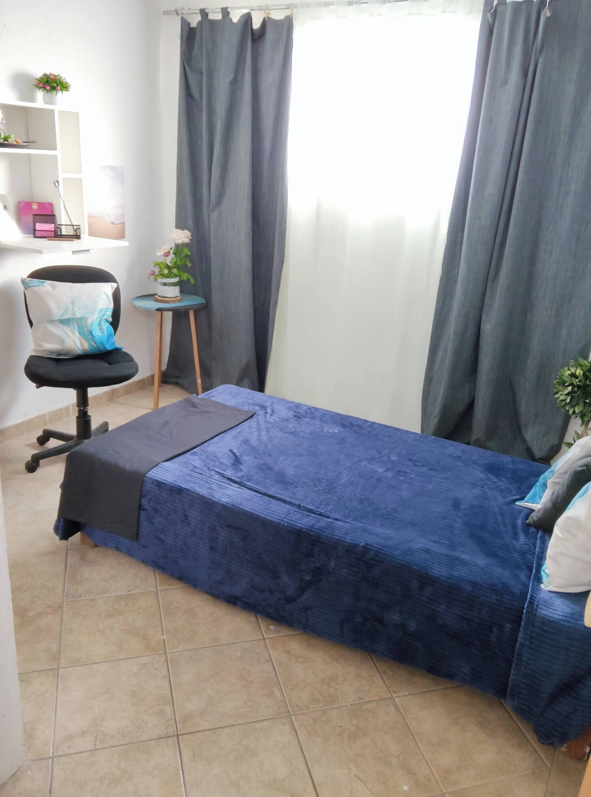 Una habitación pequeña y ordenada con una cama individual, una silla, una mesita y un estante con objetos personales.