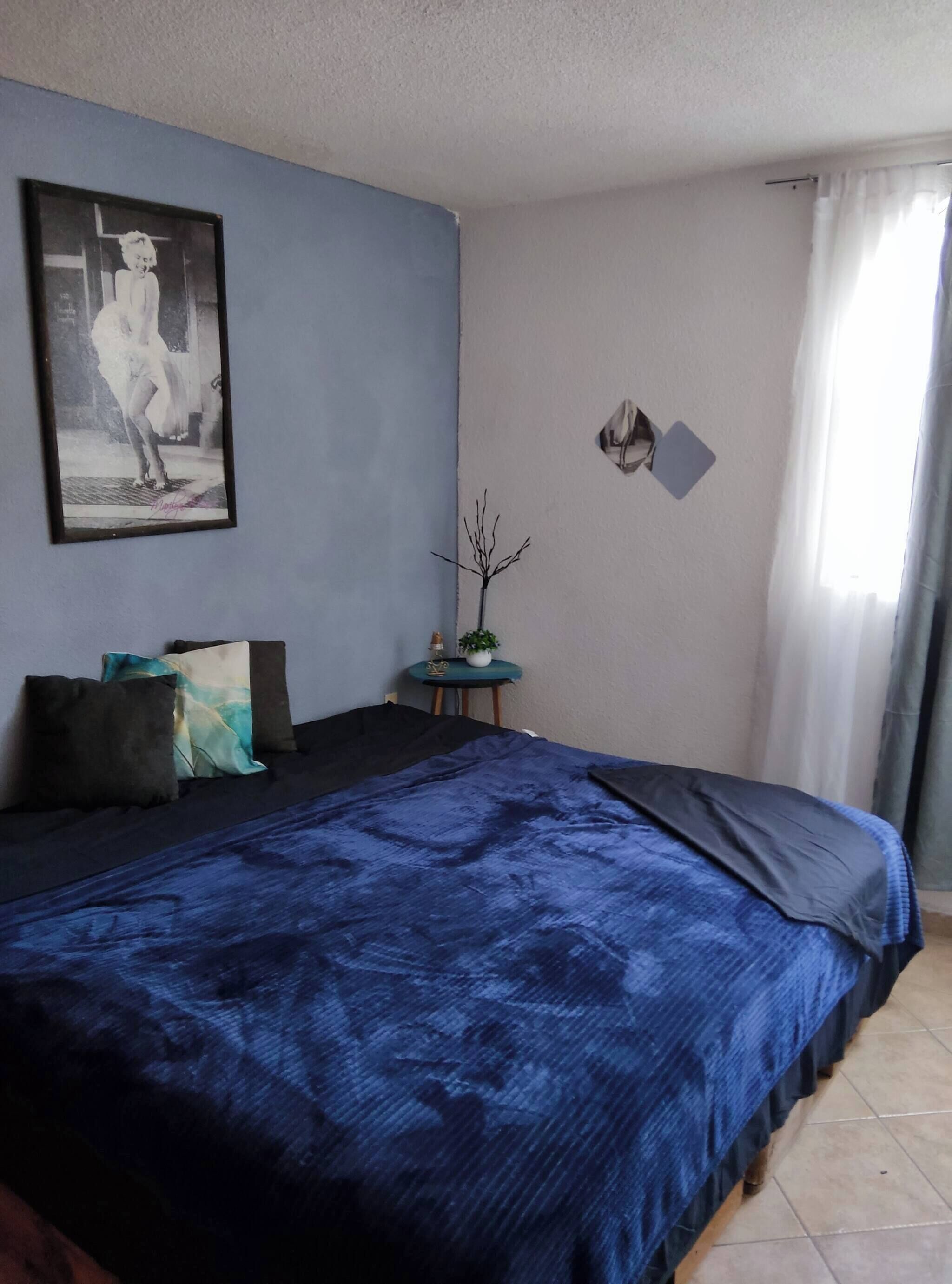 A bedroom with a large bed, a small table with a plant, a framed picture on the wall, and a window with curtains.