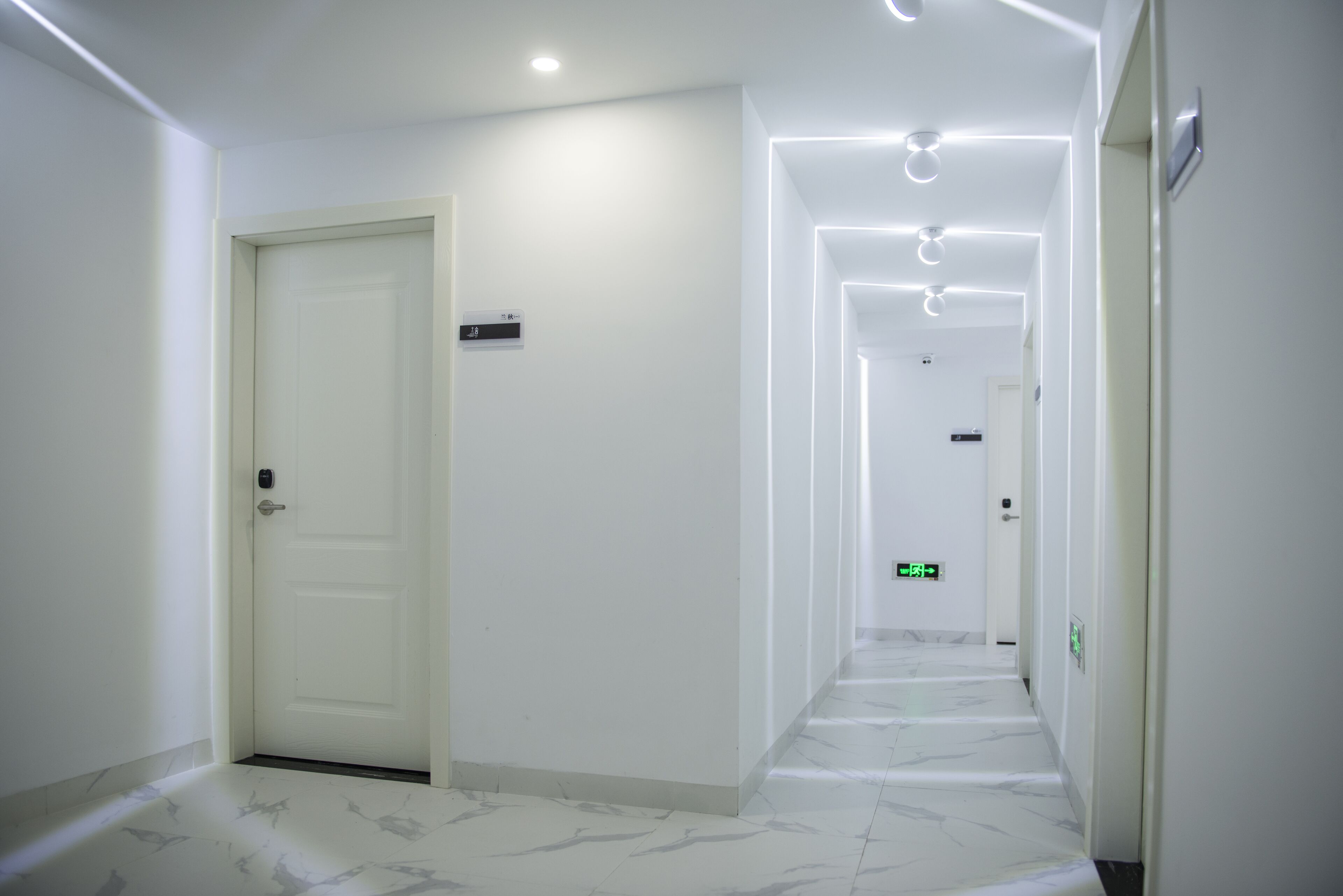 A clean, modern hallway with white walls and marble flooring, leading to multiple doors and an emergency exit sign.