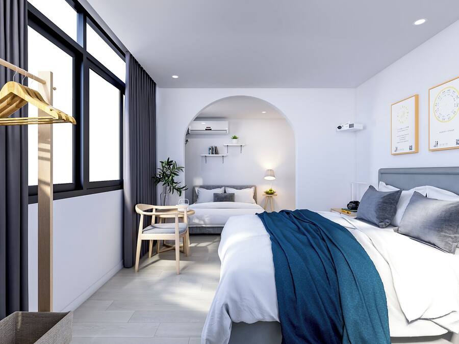 A modern hotel room with a bed, a desk, and a chair. There is a window with curtains, a plant, and a lamp.