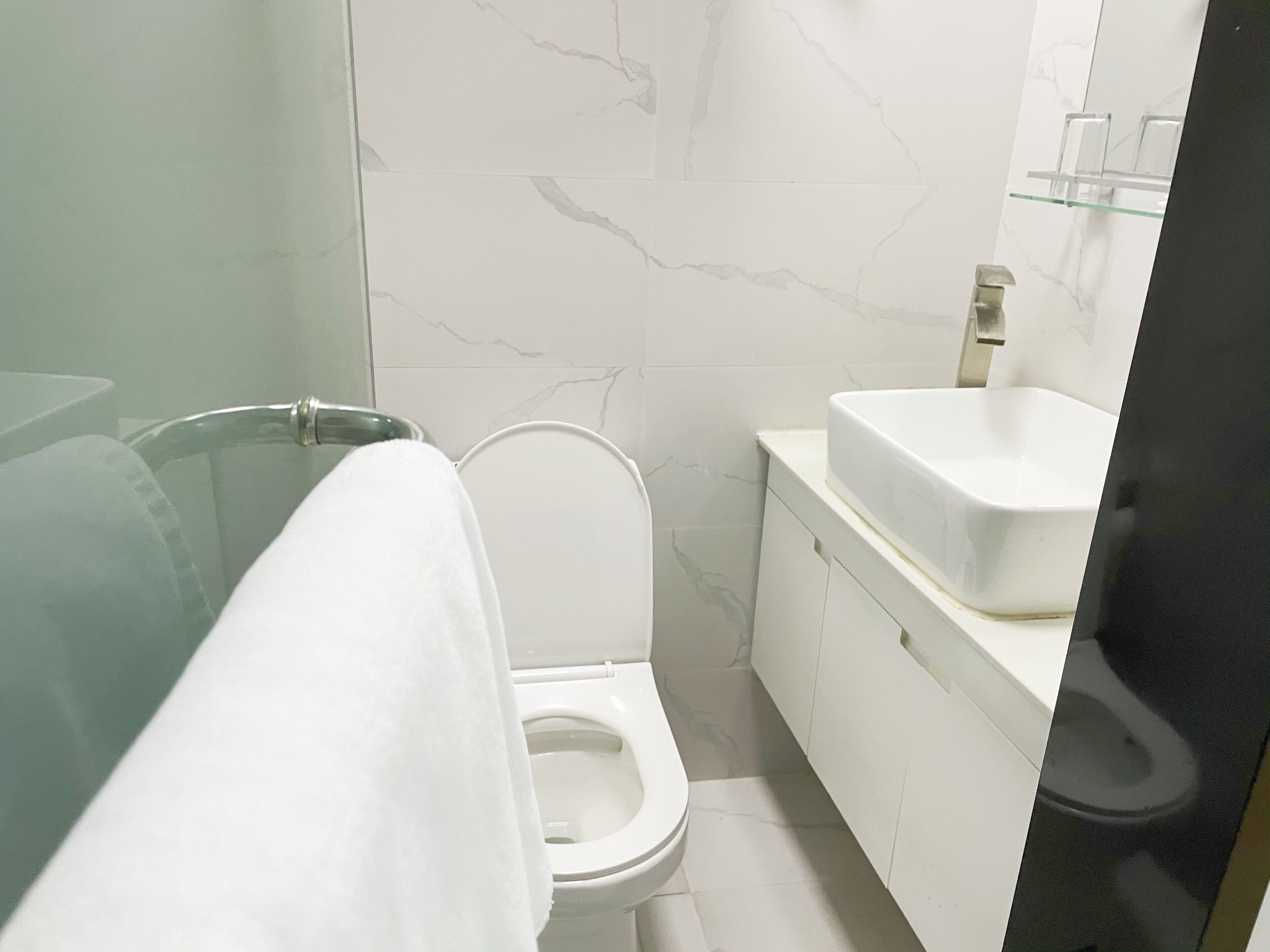 Un baño con cabina de ducha de vidrio, inodoro blanco y lavamanos blanco con resalte de mármol.