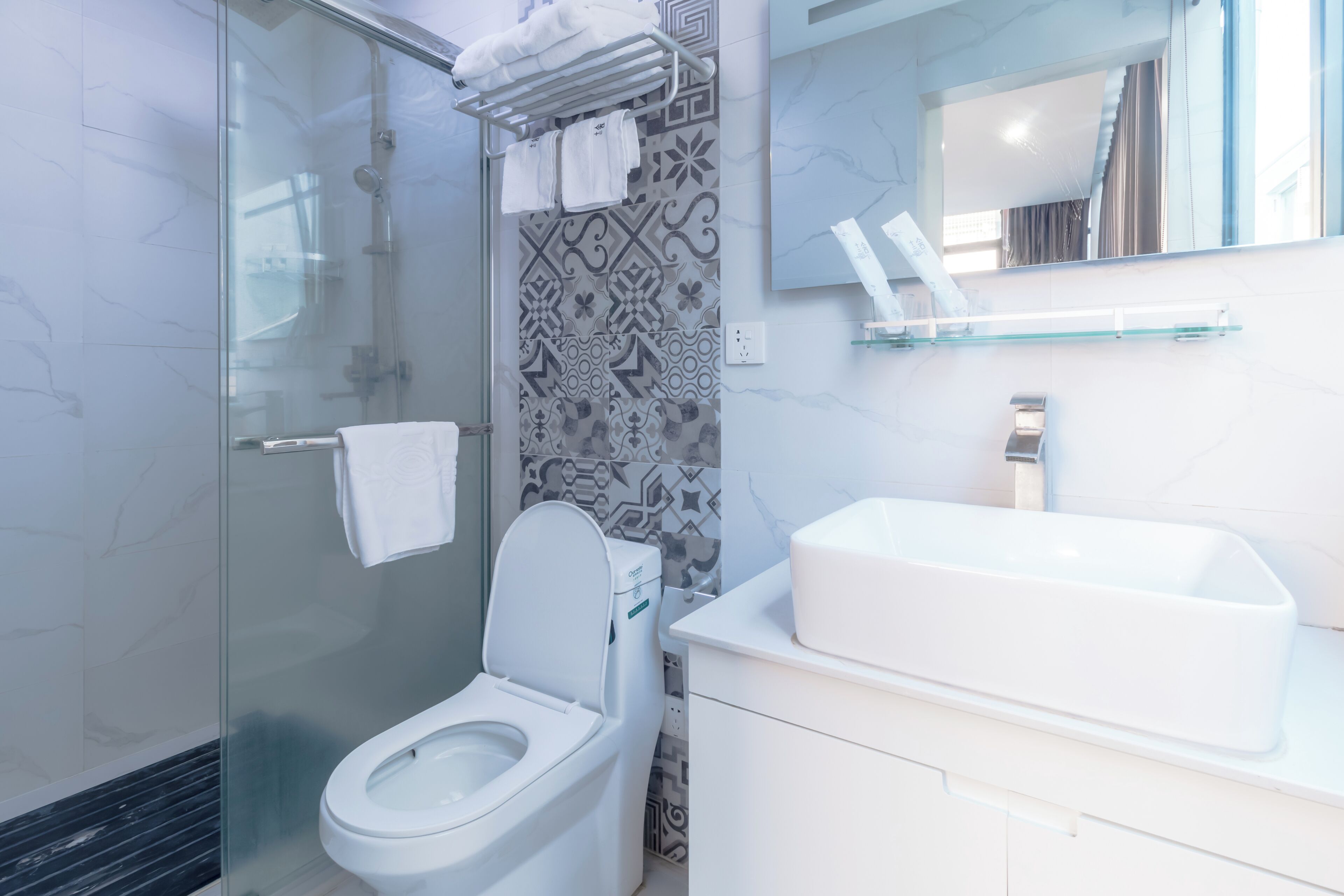 Un baño moderno con ducha de vidrio, inodoro blanco, lavamanos grande de color blanco y paredes con diseño de azulejos.