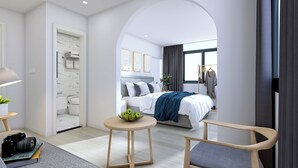 Ein modernes Hotelzimmer mit einem Bett, einem runden Holztisch mit Obstschale, einem Sessel und einem Schreibtisch mit Kamera.