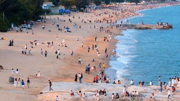 Ein belebter Strand mit Menschen, die auf dem Sand und den Felsen entlanggehen und sitzen.