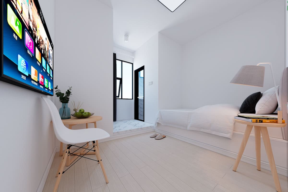Ein modernes, minimalistisches Schlafzimmer mit einem großen Flachbildfernseher, einem weißen Sofa, einem Nachttisch mit Büchern und einer kleinen Pflanze.