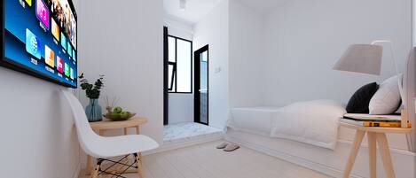 A modern, minimalist bedroom with a large flat-screen TV, a white sofa, a bedside table with books, and a small plant.