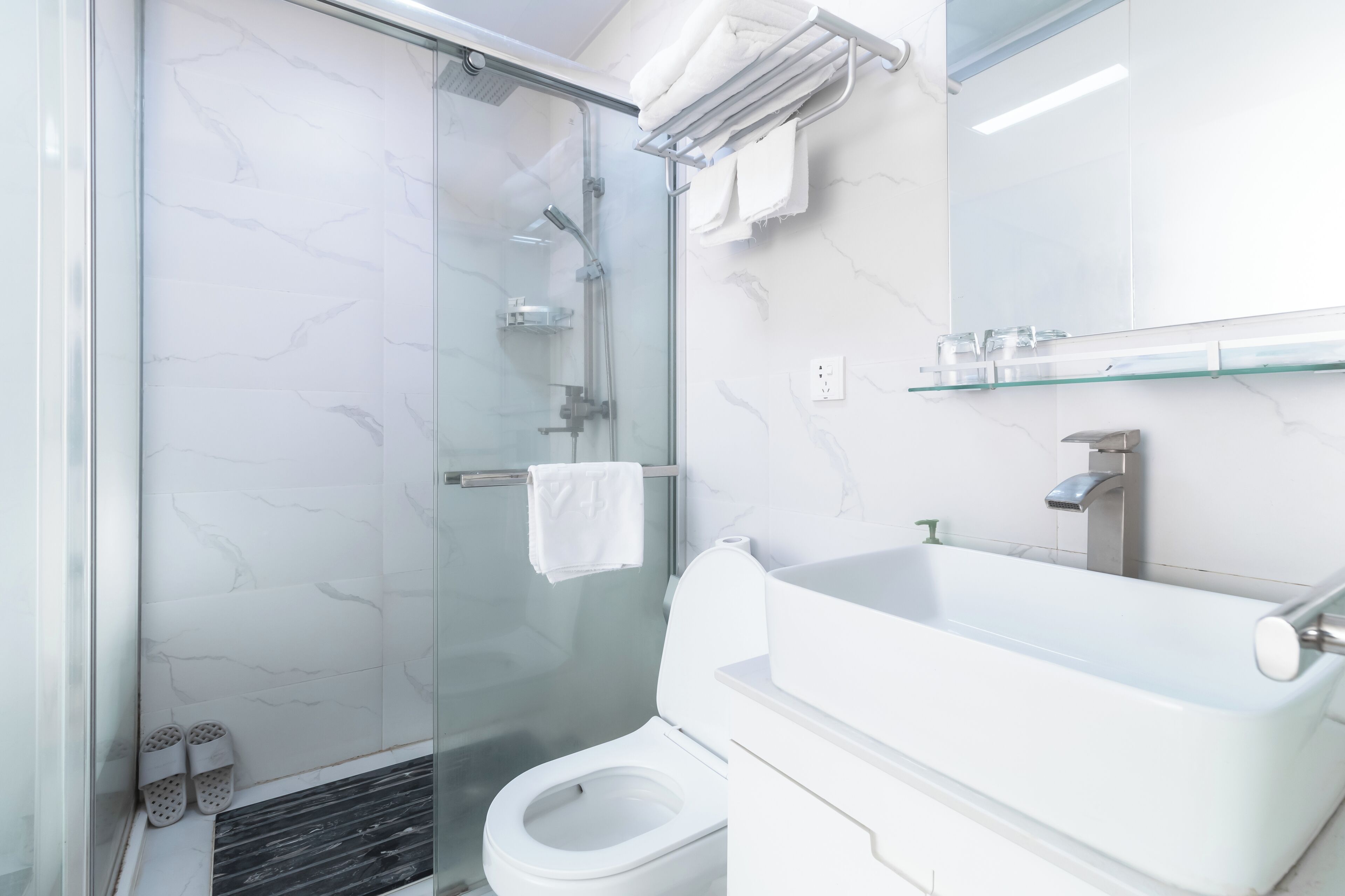 Une salle de bain moderne avec une douche à parois de verre, des toilettes blanches et un lavabo surmonté d’une plaque de verre.