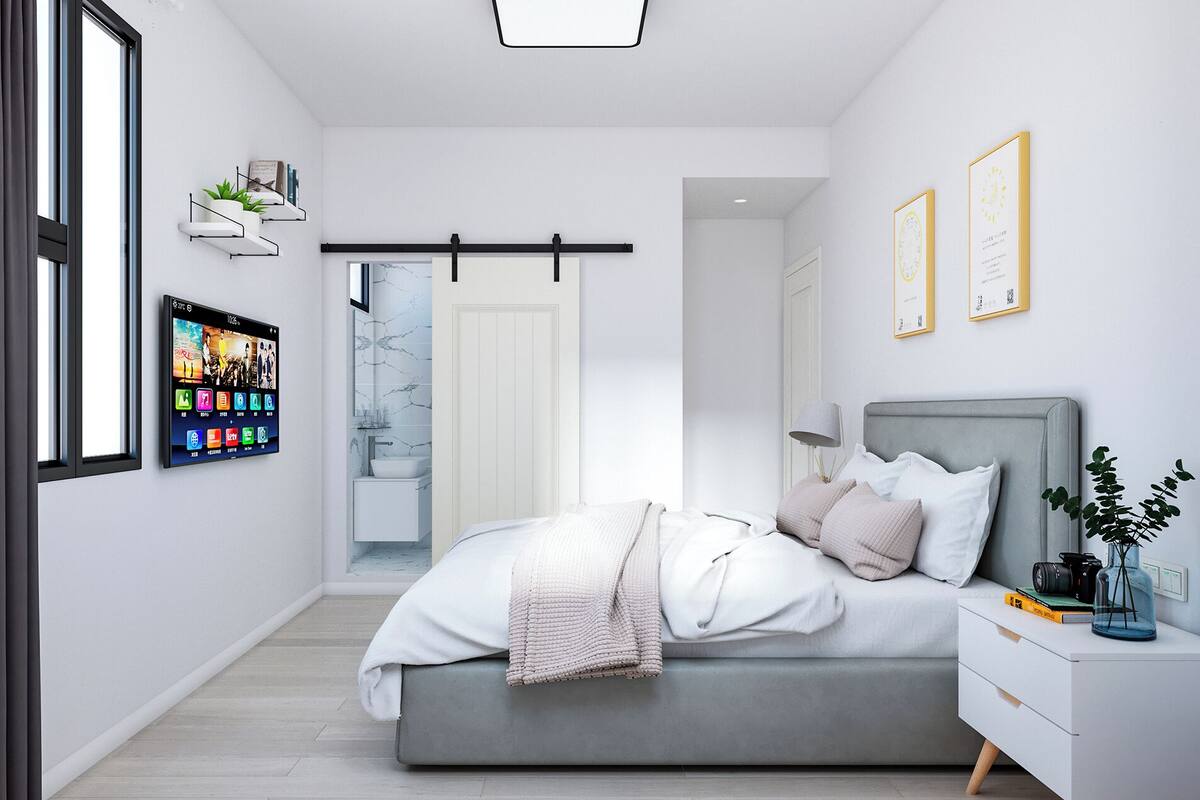 Ein modernes Schlafzimmer mit grauem Bett, weißen Wänden, einem an der Wand befestigten Fernseher und einem Nachttisch mit Lampe und Büchern.