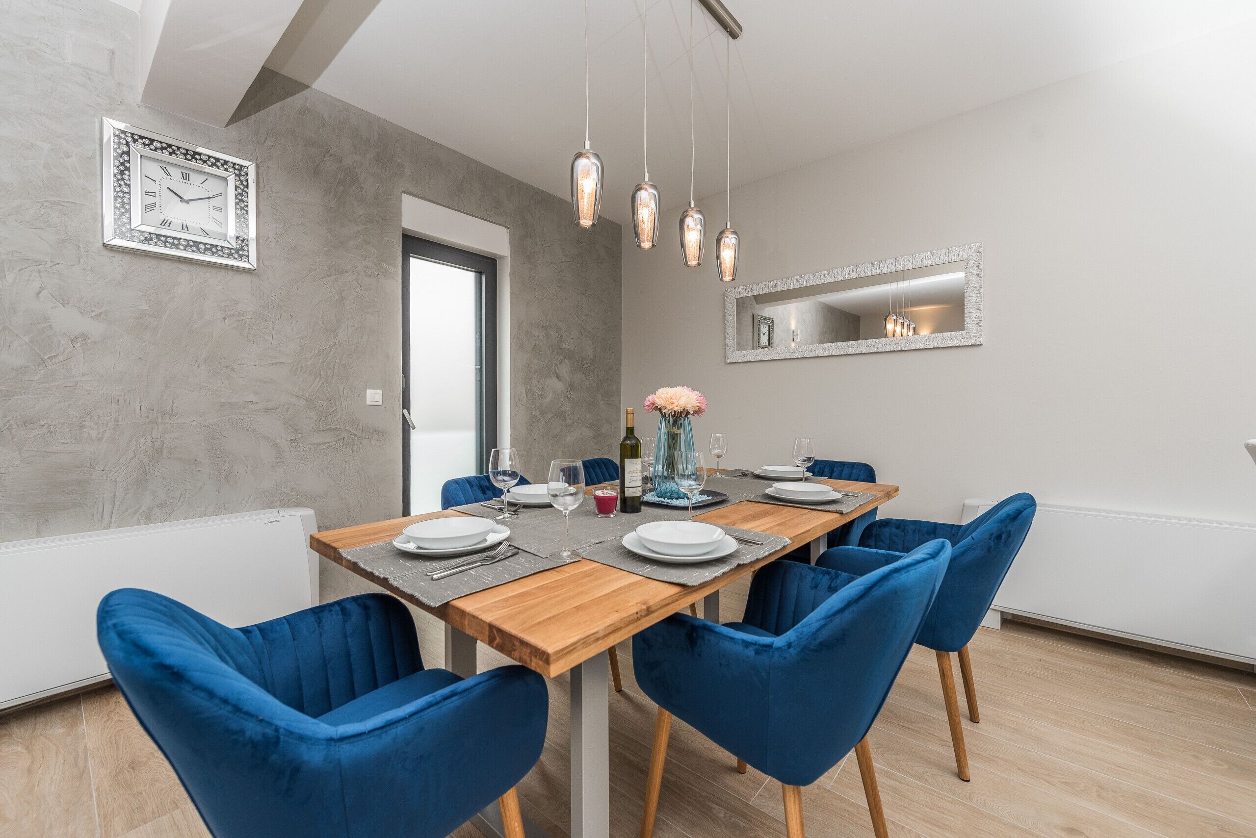 A modern dining room with a wooden table set for six, blue chairs, hanging pendant lights, a wall clock, and a mirror above the table.