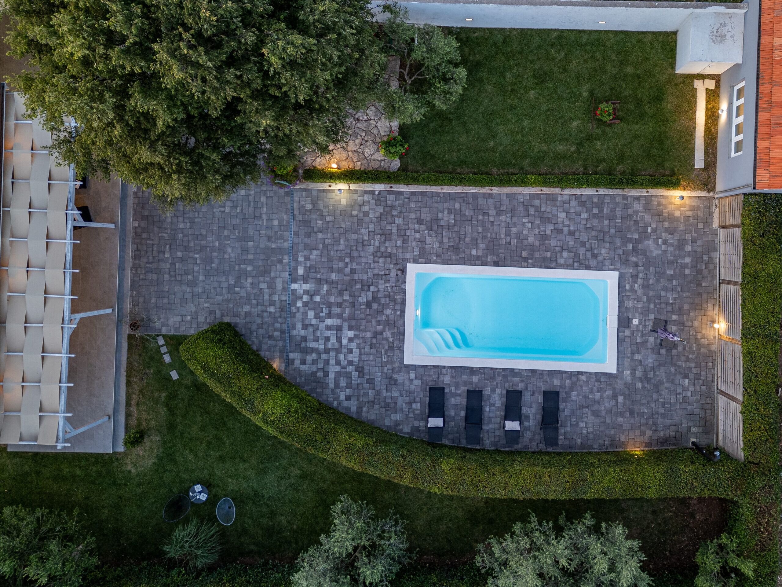Vue aérienne d’une cour arrière avec une piscine rectangulaire, un patio dallé et un espace détente comprenant des chaises longues et une table.