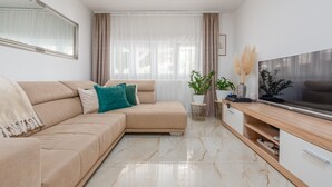Una sala de estar moderna con un sofá color beige, un televisor de pantalla plana, piso de mármol y un ventanal con cortinas de gasa.