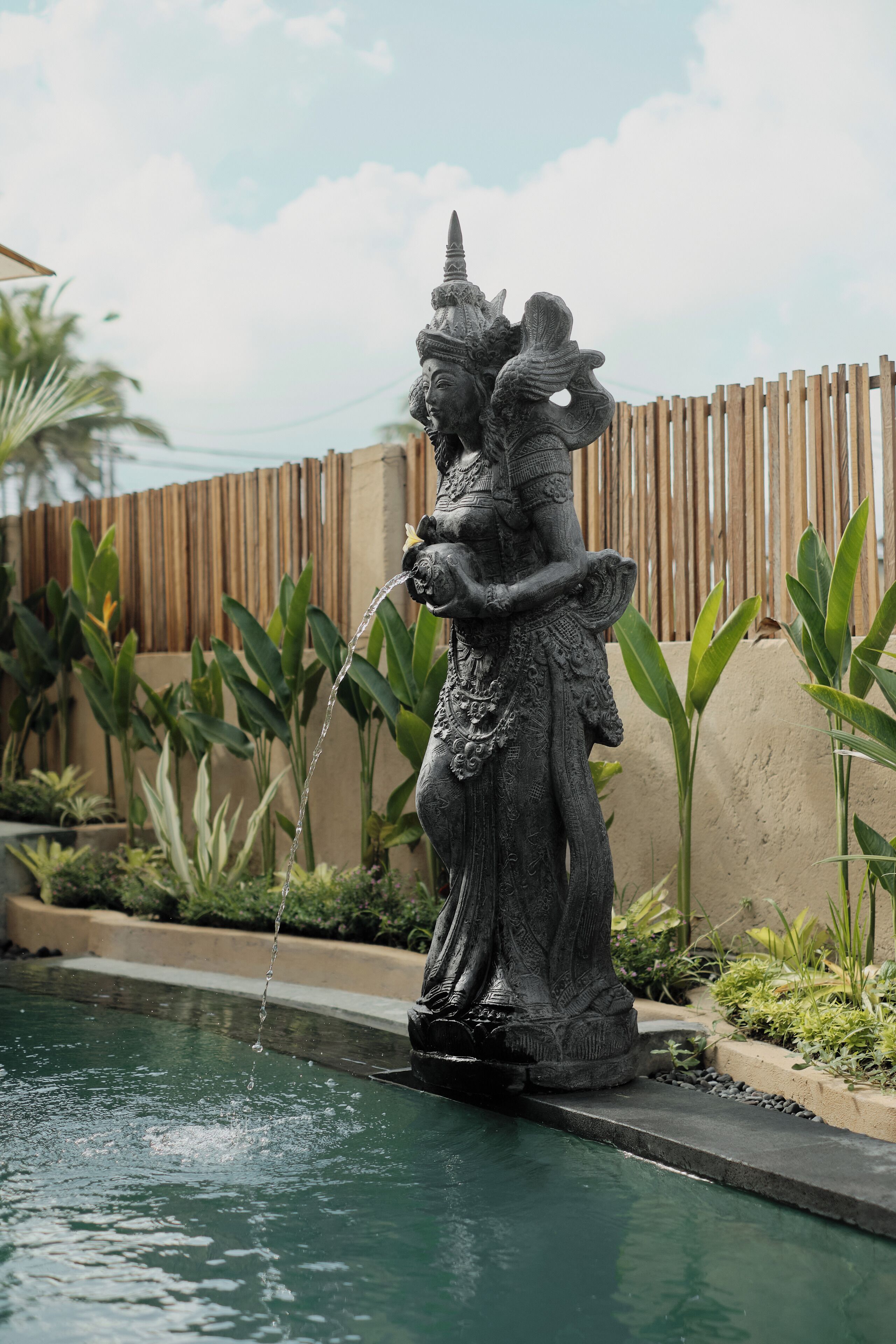 Eine Statue neben einem Pool mit Holzzaun und tropischer Bepflanzung im Hintergrund.