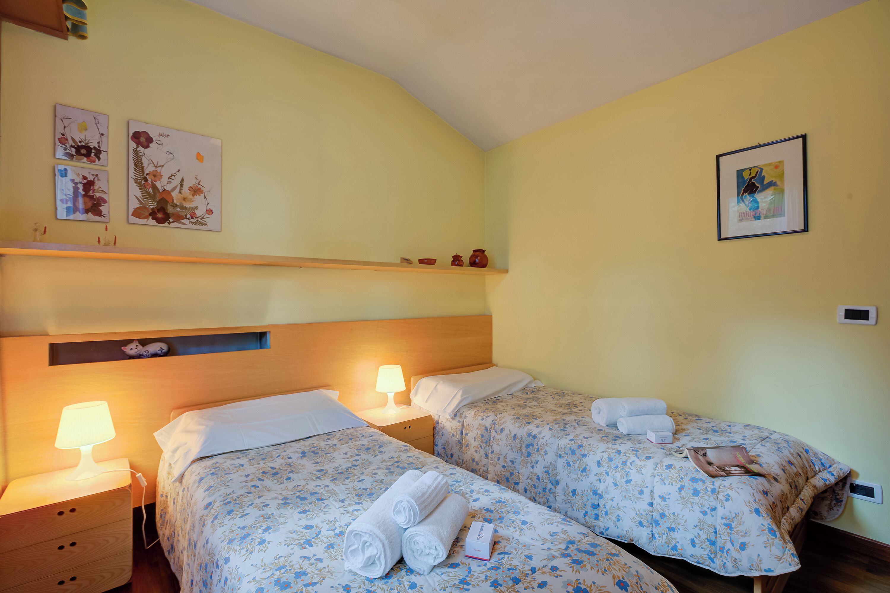 A hotel room with two beds, each with floral bedding and white pillows. There are bedside tables with lamps, a shelf with decorative items, and artwork on the walls.