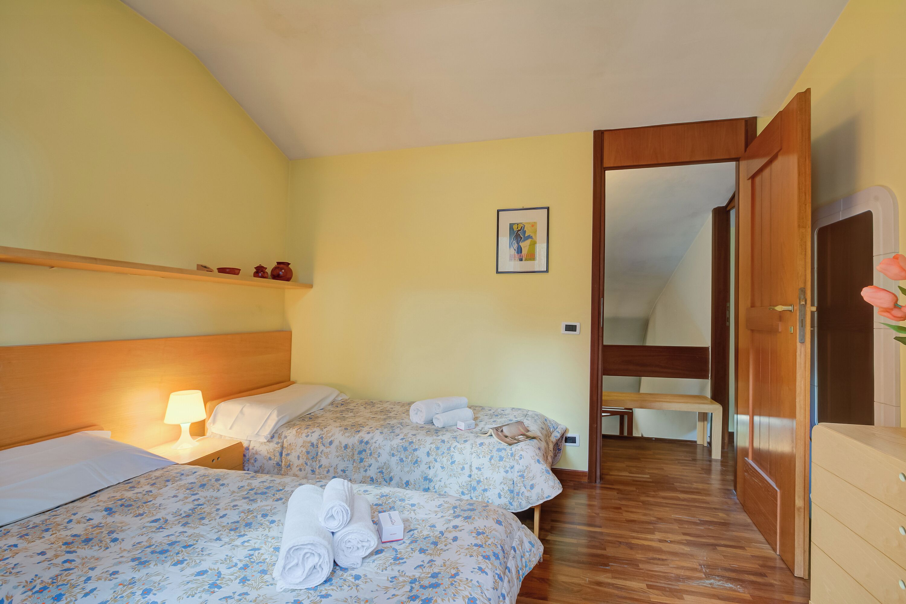 A hotel room with two beds, a wooden headboard, a bedside table with a lamp, a bench, and a wooden wardrobe.