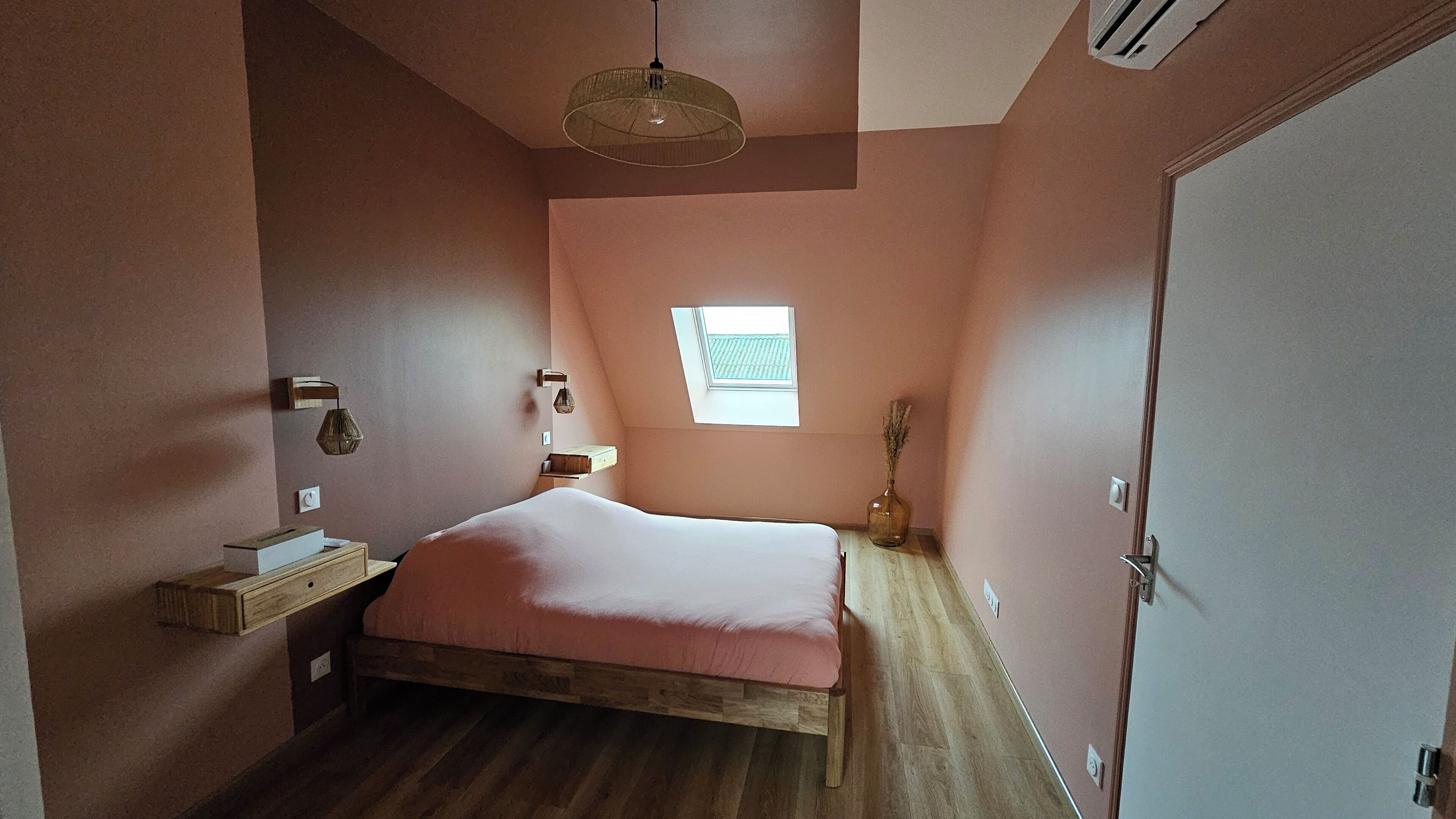 A bedroom with a wooden bed, a skylight, a bedside table, and a vase with dried flowers.