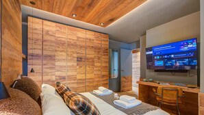 Una camera d'albergo moderna con soffitto in legno, un letto con lenzuola bianche, una scrivania con una sedia e una televisione che mostra l'interfaccia di un servizio di streaming.