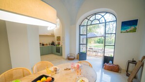 A dining area with a table set for four, a fruit bowl, and a view of the garden through a large window. - Villa Vallone Luxury With Pool (Galatina)