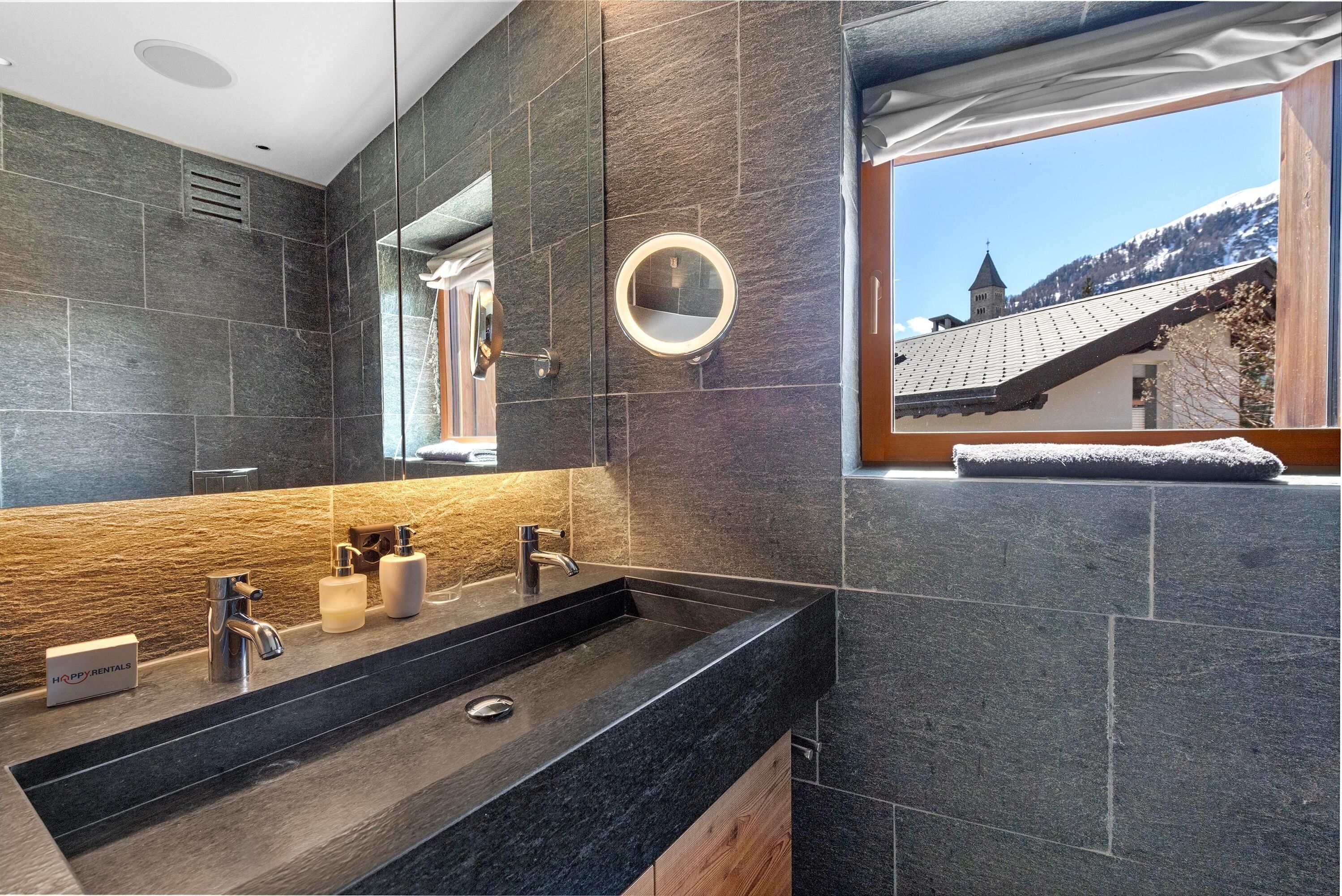 Een moderne badkamer met een grote spiegel, dubbele wastafels en uitzicht op een berg en kerk door een raam.