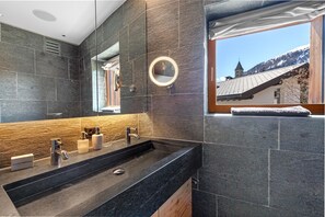 A modern bathroom with a large mirror, dual sinks, and a view of a mountain and church through a window. - Maison Montagne in Samedan (Samedan)