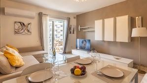 Ein modernes Wohnzimmer mit einer Couch, einem Esstisch gedeckt mit Tellern und Gläsern, einem Fernseher und einem Balkon mit Vorhängen.