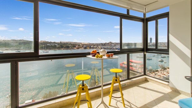 A room with large windows overlooking a marina and cityscape. There are two yellow stools and a small table with fruits and a teapot. - Magnificent Sunny Seafront Apart Sliema (Sliema)