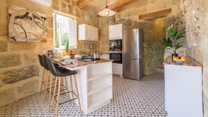 Cocina rústica con paredes de piedra, techo de madera y piso de baldosas. Cuenta con una isla central con taburetes, armarios blancos, un refrigerador de acero inoxidable y una lámpara colgante.
