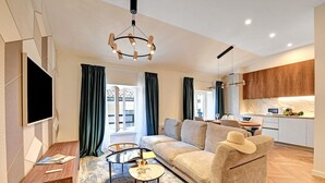 A modern living room with a sofa, coffee table, and TV. There is a kitchen area with cabinets and a countertop. - Novos Diamond in Lugano (Lugano)
