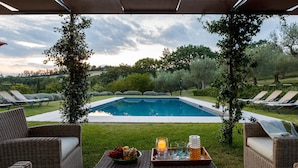 A poolside area with lounge chairs, a wicker sofa, a tray with fruit and drinks, and a view of the pool and surrounding greenery. - Casa Eugenia 8 Emma Villas (Pesaro)