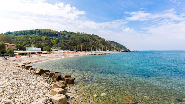 A pebble beach with clear water, a rocky shoreline, and a hillside with buildings in the background. - Casa Eugenia 8 Emma Villas (Pesaro)