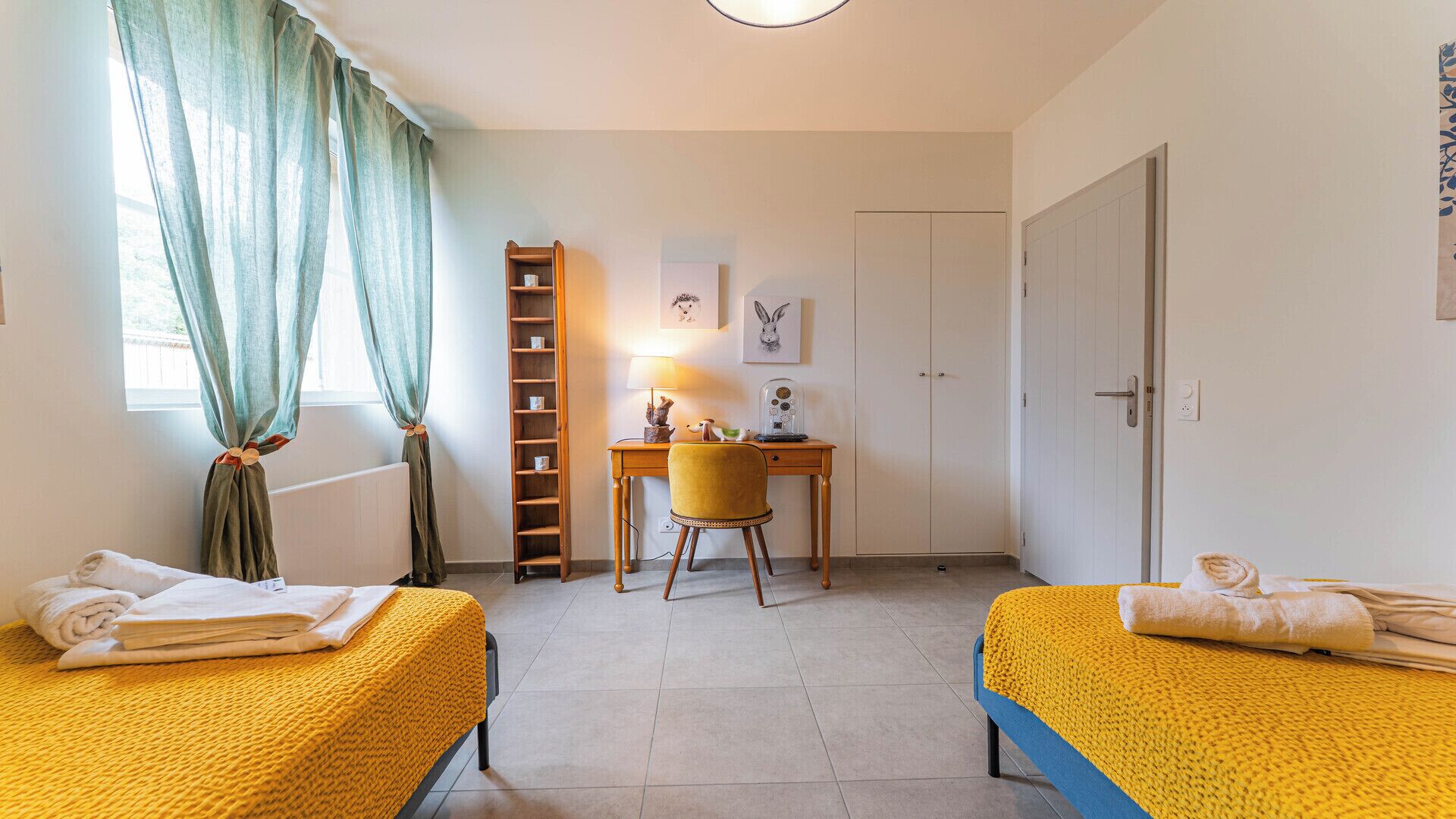 A room with two beds, a desk, a chair, a wardrobe, and a window with curtains.