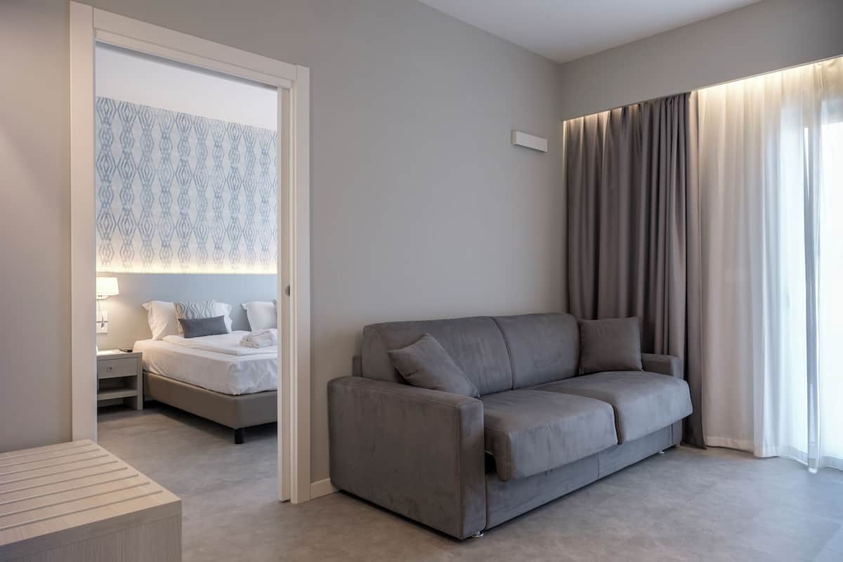 A modern hotel room with a grey sofa, a bed with white linens, a bedside table, and a large window with curtains.