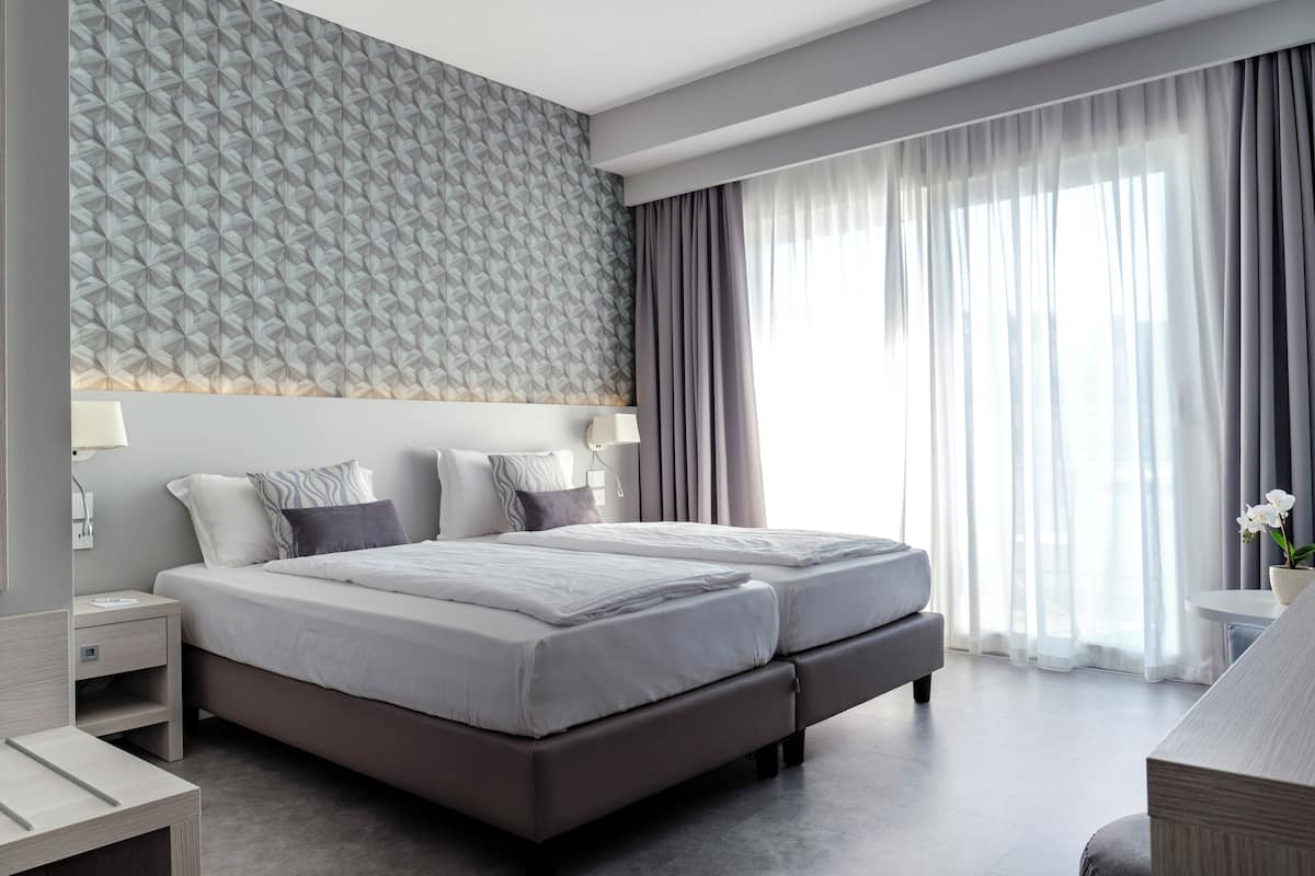 A modern hotel room with a large bed, a bedside table, a lamp, and a view of the outdoors through sheer curtains.