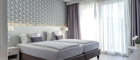 A modern hotel room with a large bed, a bedside table, a lamp, and a view of the outdoors through sheer curtains.