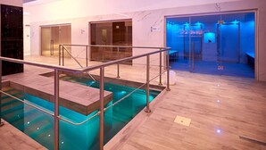 Un área de piscina interior moderna con barandilla de vidrio y pasamanos de metal, paredes de mármol y suelo de madera.