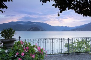 A lakeside view with mountains in the background, a metal railing, and potted plants with flowers. - Residenza Menaggio - Mimosa (Menaggio)