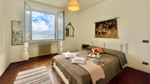 Un dormitorio con un ventanal grande, una cama con cubrecama marrón, una mesita de noche con lámpara y un cuadro en la pared.