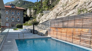 En swimmingpool med en mur av sten och träpanel, omgiven av ett bergigt landskap och en byggnad med välvda fönster.