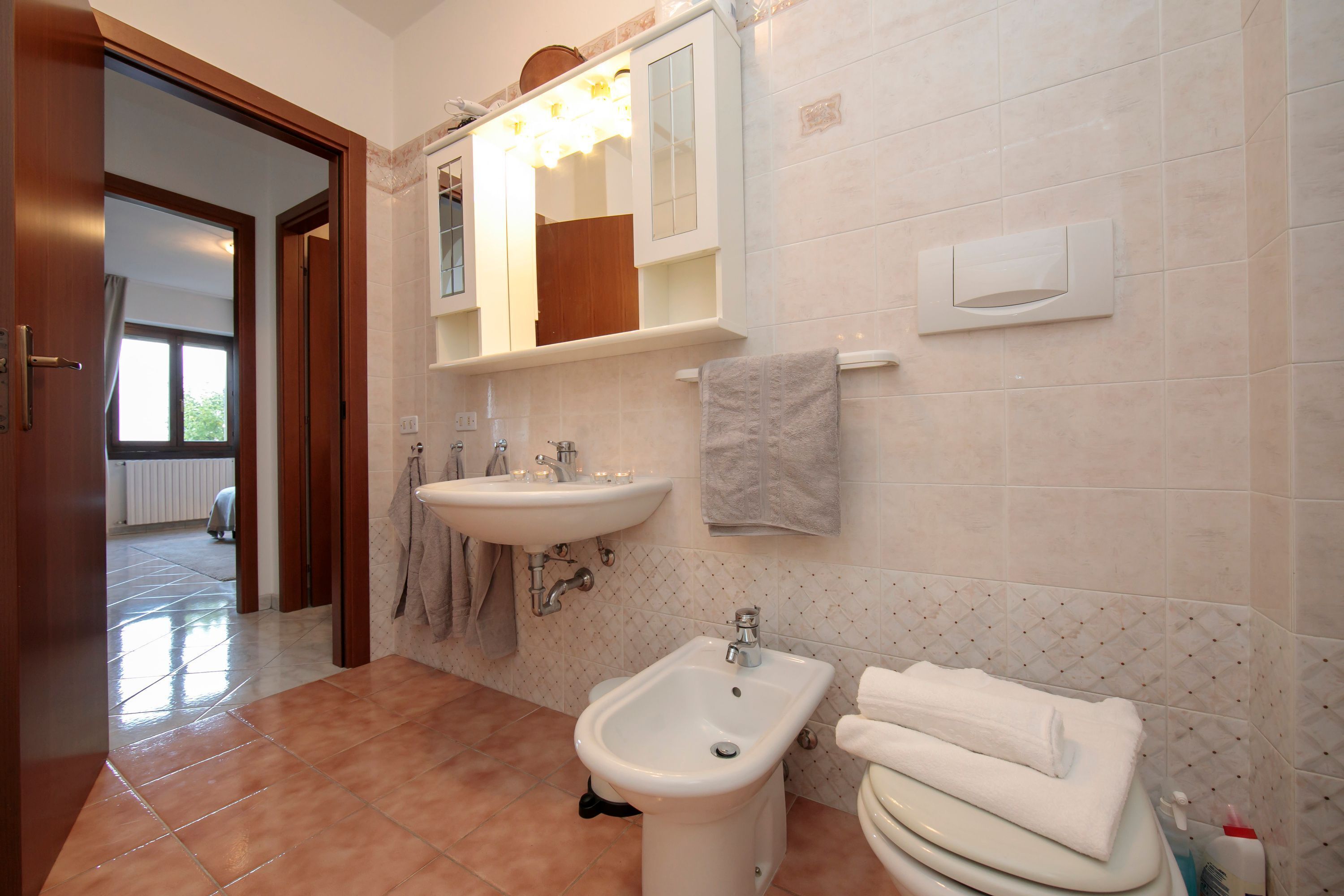 Une salle de bain équipée d’un bidet, d’un lavabo et d’un miroir. On y trouve également un porte-serviettes avec des serviettes et une fenêtre avec des rideaux.