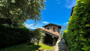 A stone house with a red-tiled roof, surrounded by lush greenery and a stone pathway leading to the entrance. - Villetta Alessia in Menaggio (San Siro)